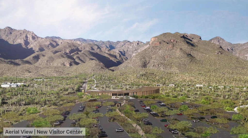 An aerial view shows the Sabino Canyon Visitor Center, a brown, curved building, surrounded by a parking lot filled with cars. The landscape includes desert vegetation and is set against a backdrop of mountains under a clear sky.