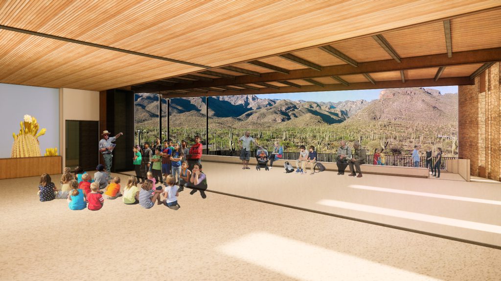 Bright visitor center educational space with high ceiling, 180° mountain view. Presenter and children gathered, engaging atmosphere.